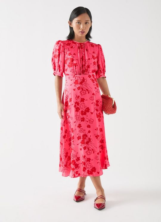 A woman wearing a pink floral tea dress with puffed short sleeves, a crew neck, and a fitted waist, paired with red heels and holding a red clutch.