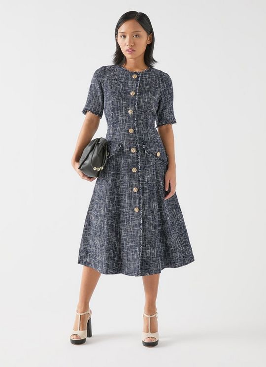 A woman wearing a midi dress made of indigo, black, and cream tweed fabric, featuring a button-through front with gold buttons, short sleeves, and frayed edges.