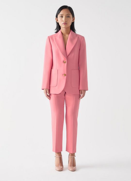 A woman wearing a pink tailored double-breasted jacket with gold buttons, matching pink pants, and nude heels, standing against a plain background.