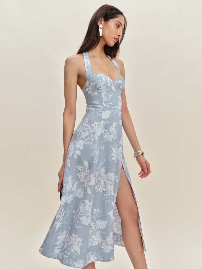 A woman wearing a light blue floral linen dress with a halter neckline, tie straps, and a front slit.