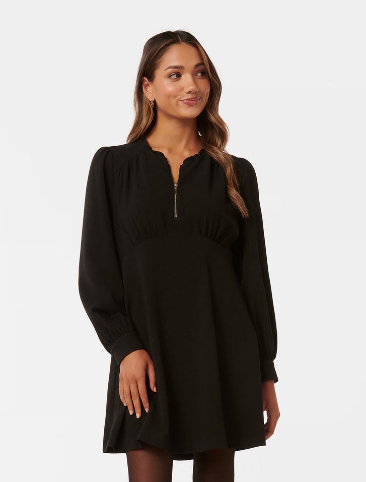 A woman wearing a black mini dress with long sleeves, a quarter zip at the neckline, and an empire mini skirt, standing against a plain background.