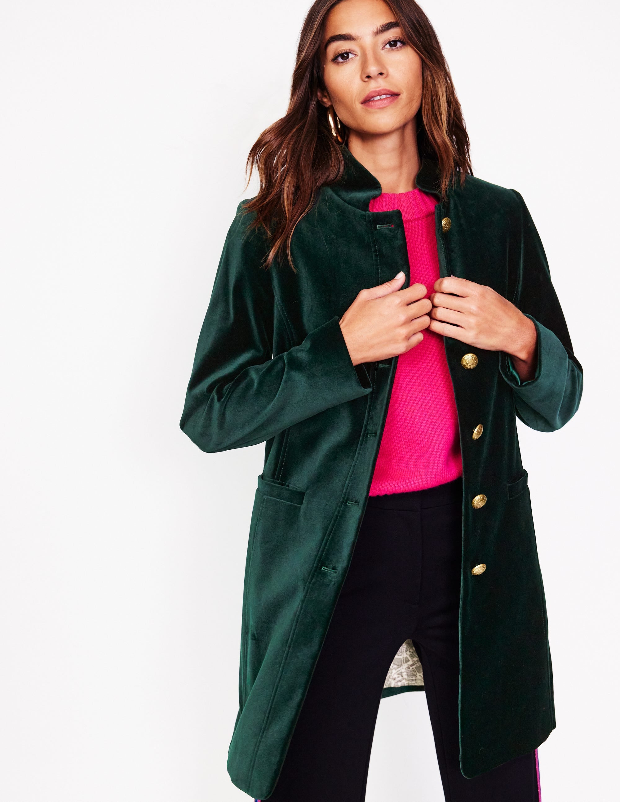 A woman wearing a dark green velvet coat with a high funnel neck, gold buttons, and welt pockets, over a bright pink top.