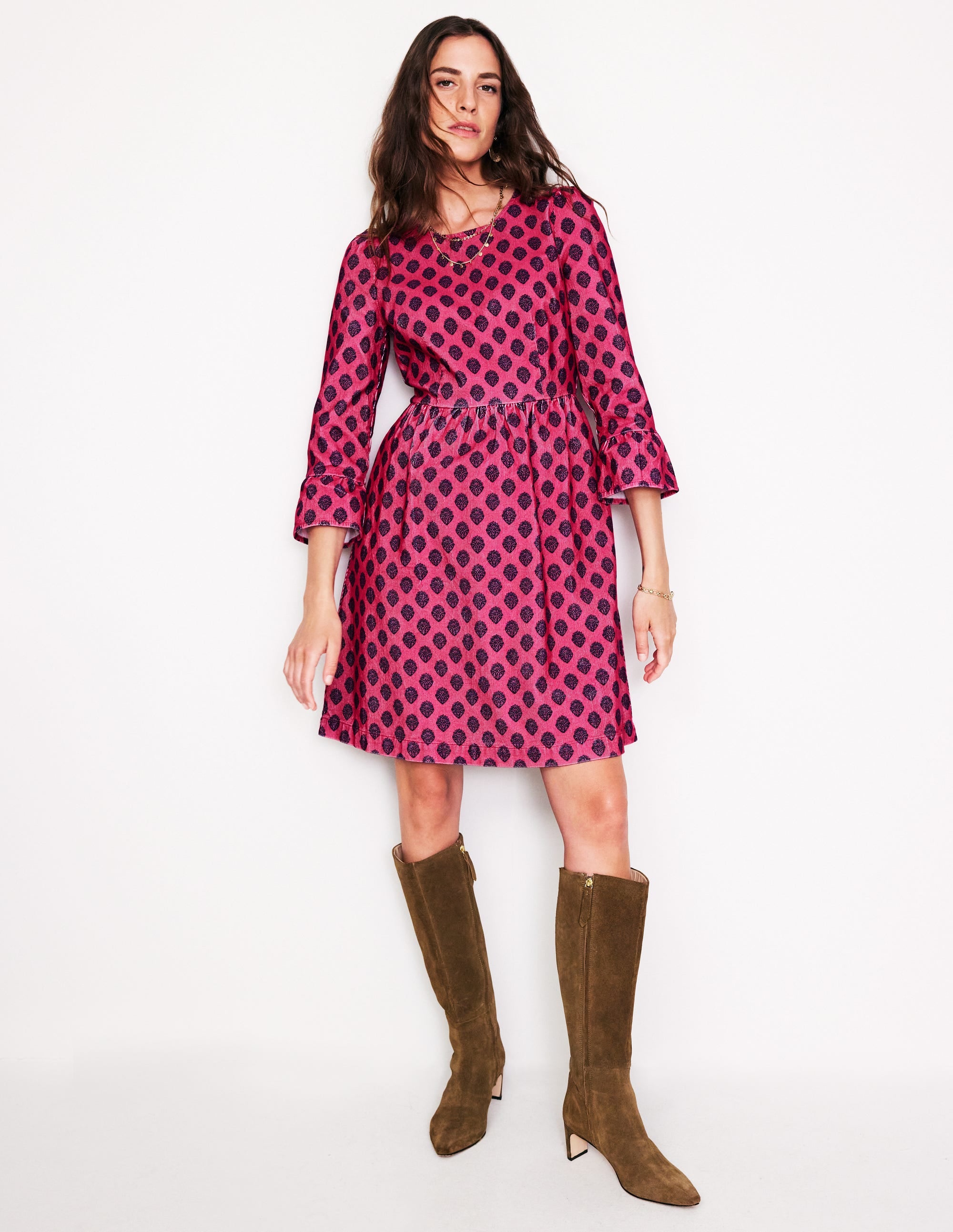 A woman wearing a pink and black patterned short cord dress with a fitted bodice and gathered skirt, paired with brown knee-high boots.