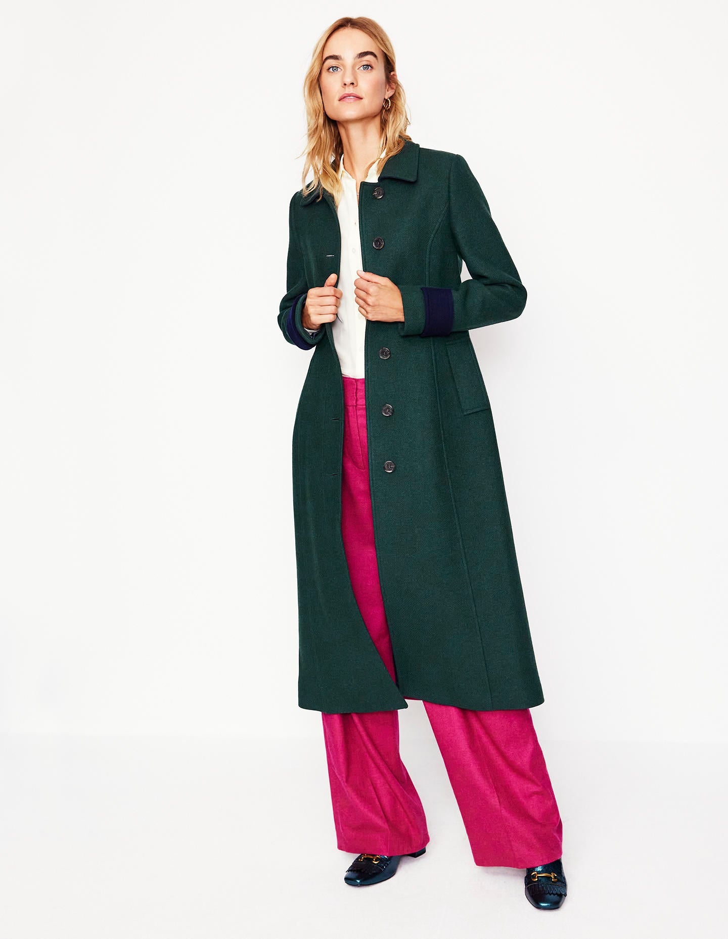A woman wearing a green colourblock midi coat with a tailored fit, princess seams, a smart collar, and functional pockets, paired with pink trousers and black shoes.