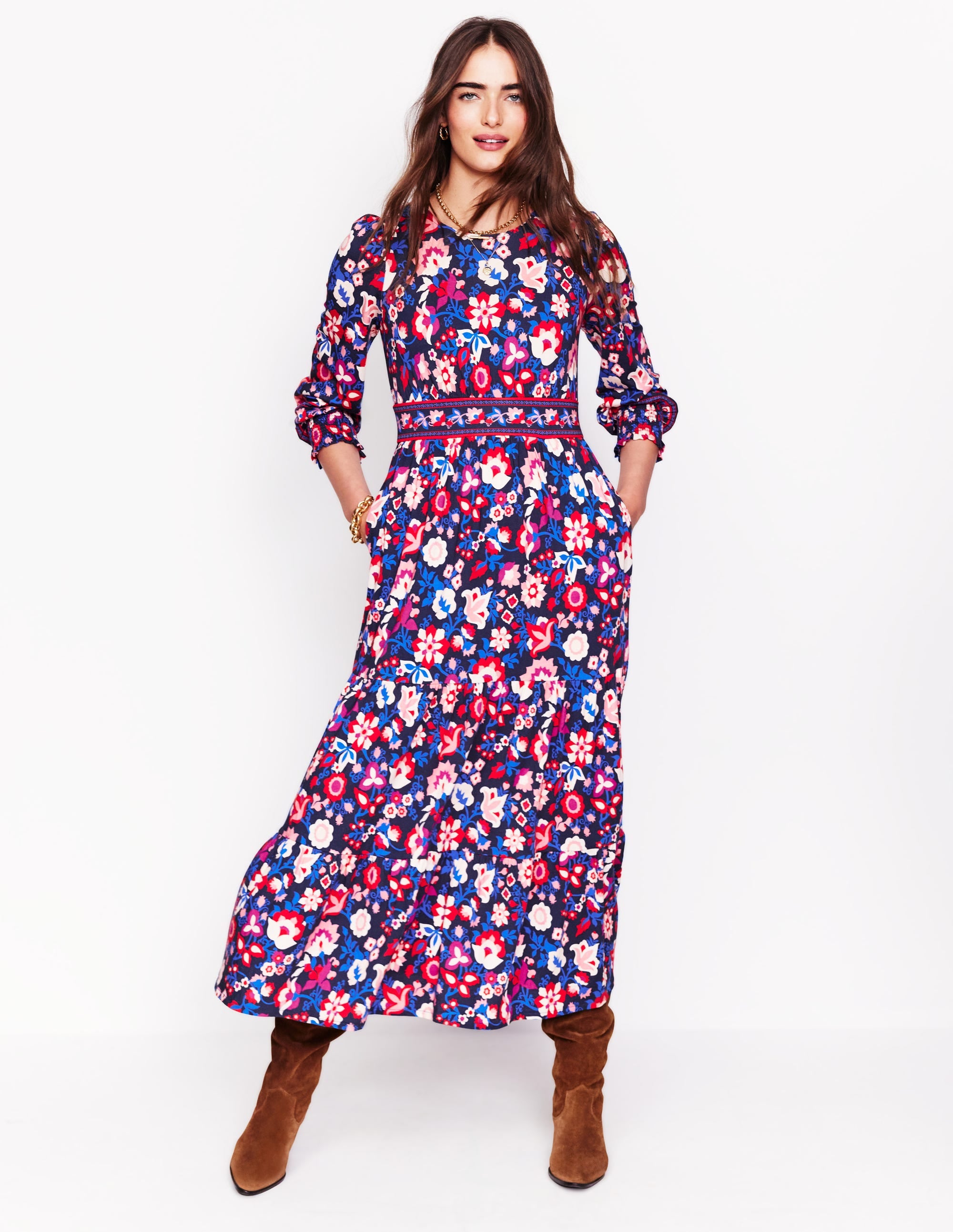 A woman wearing a long, floral patterned dress with a tiered skirt, side pockets, and blouson sleeves, in college navy with pink, white, and red flowers.