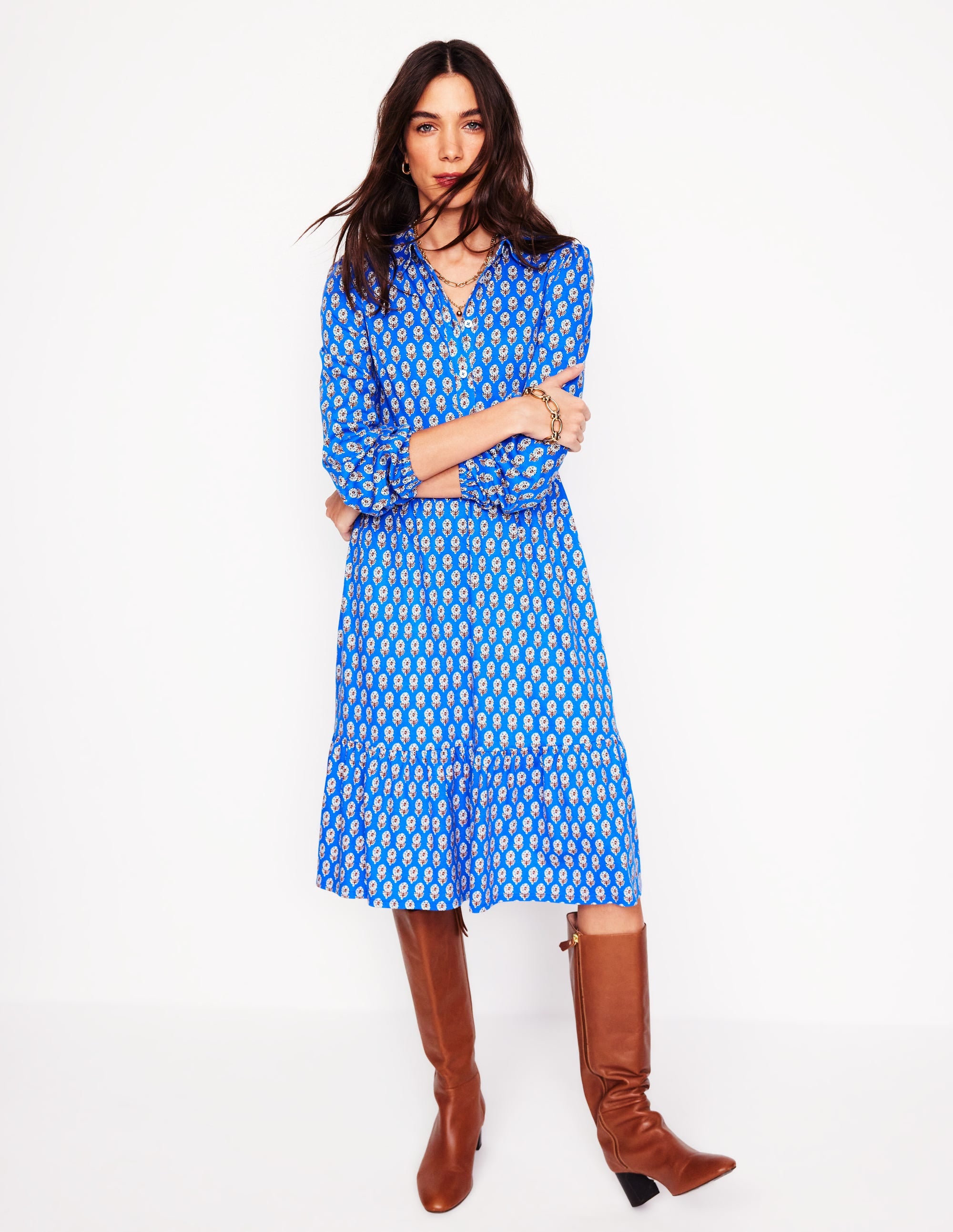 A woman wearing a blue tiered jersey dress with a floral pattern, long sleeves, collar, and side seam pockets, paired with brown knee-high boots.