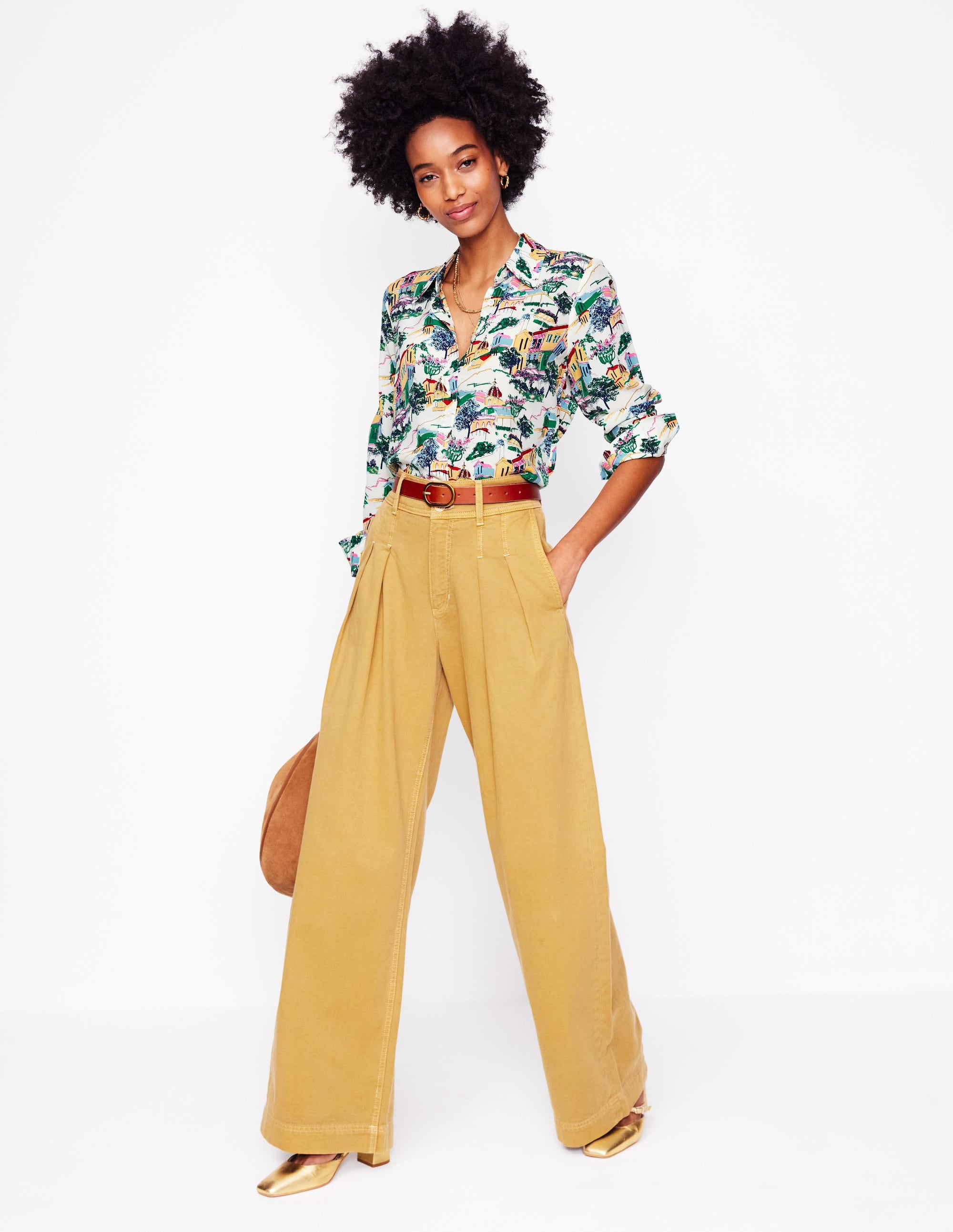 A woman wearing a multicolored painted village print silk shirt with a collar and long sleeves, tucked into high-waisted yellow trousers.