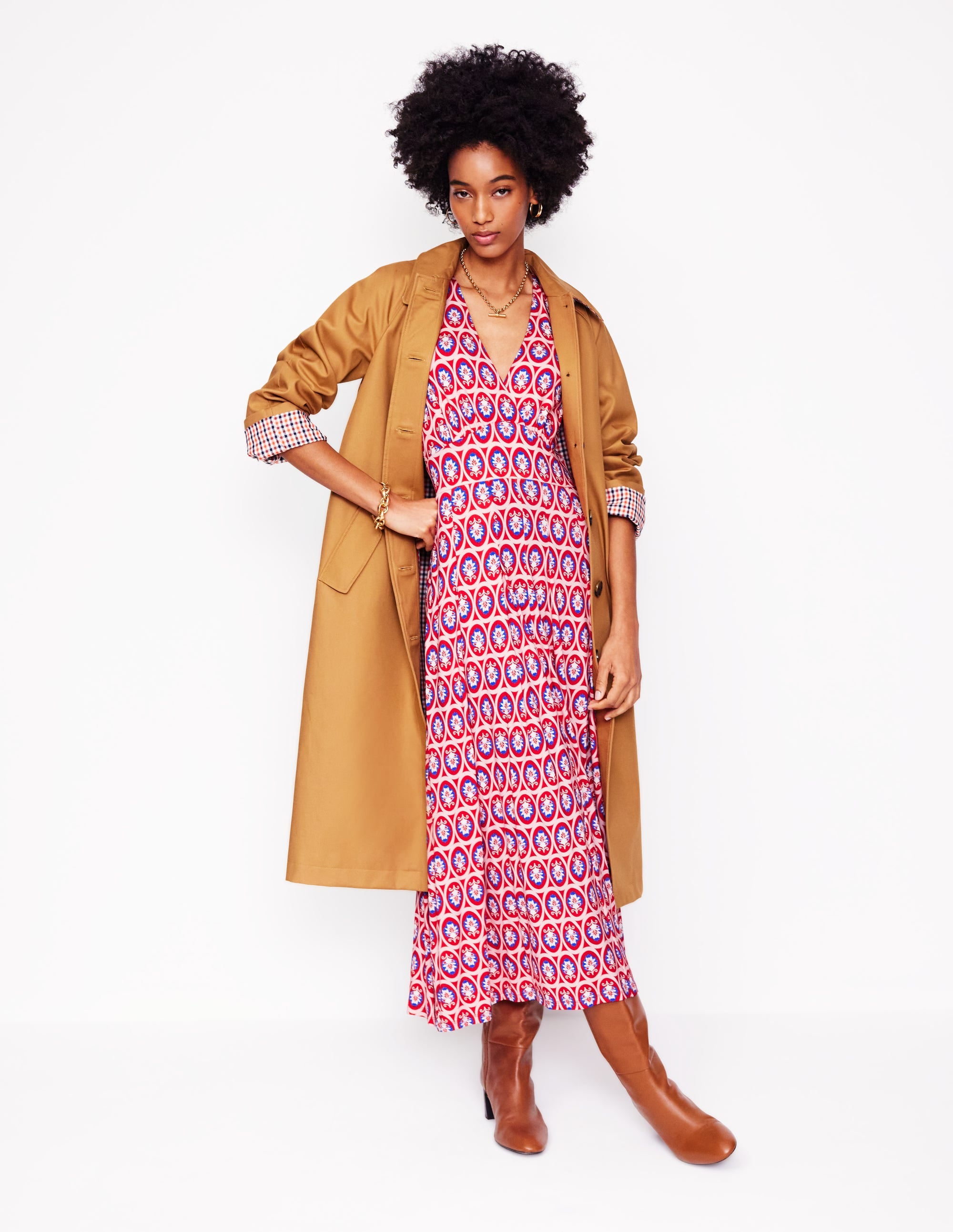 A woman wearing a red patterned pleated jersey dress with ornate blossom design, paired with a tan trench coat and brown boots.