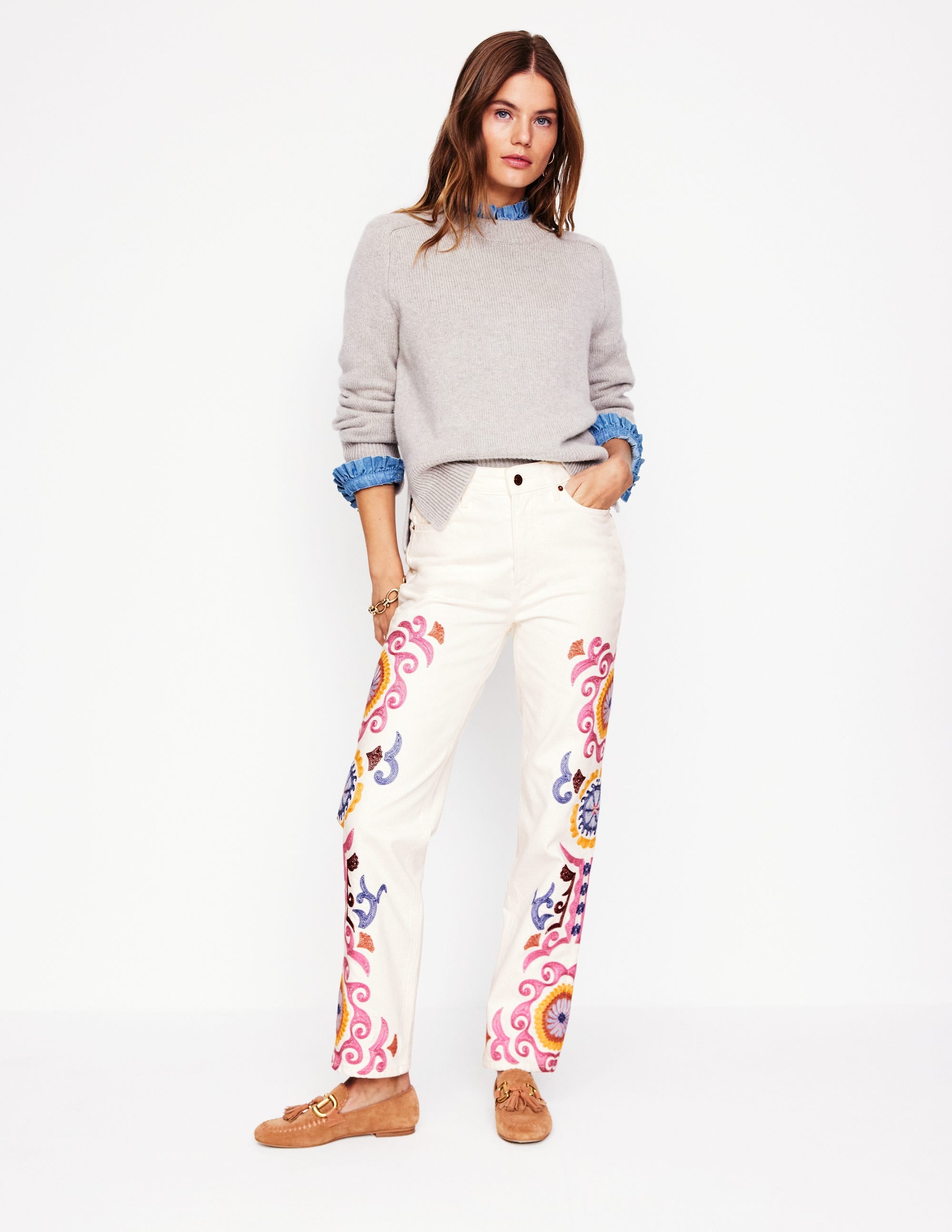 A woman wearing white high-rise embroidered jeans with colorful floral embroidery on the legs, paired with a grey sweater with blue ruffled cuffs.