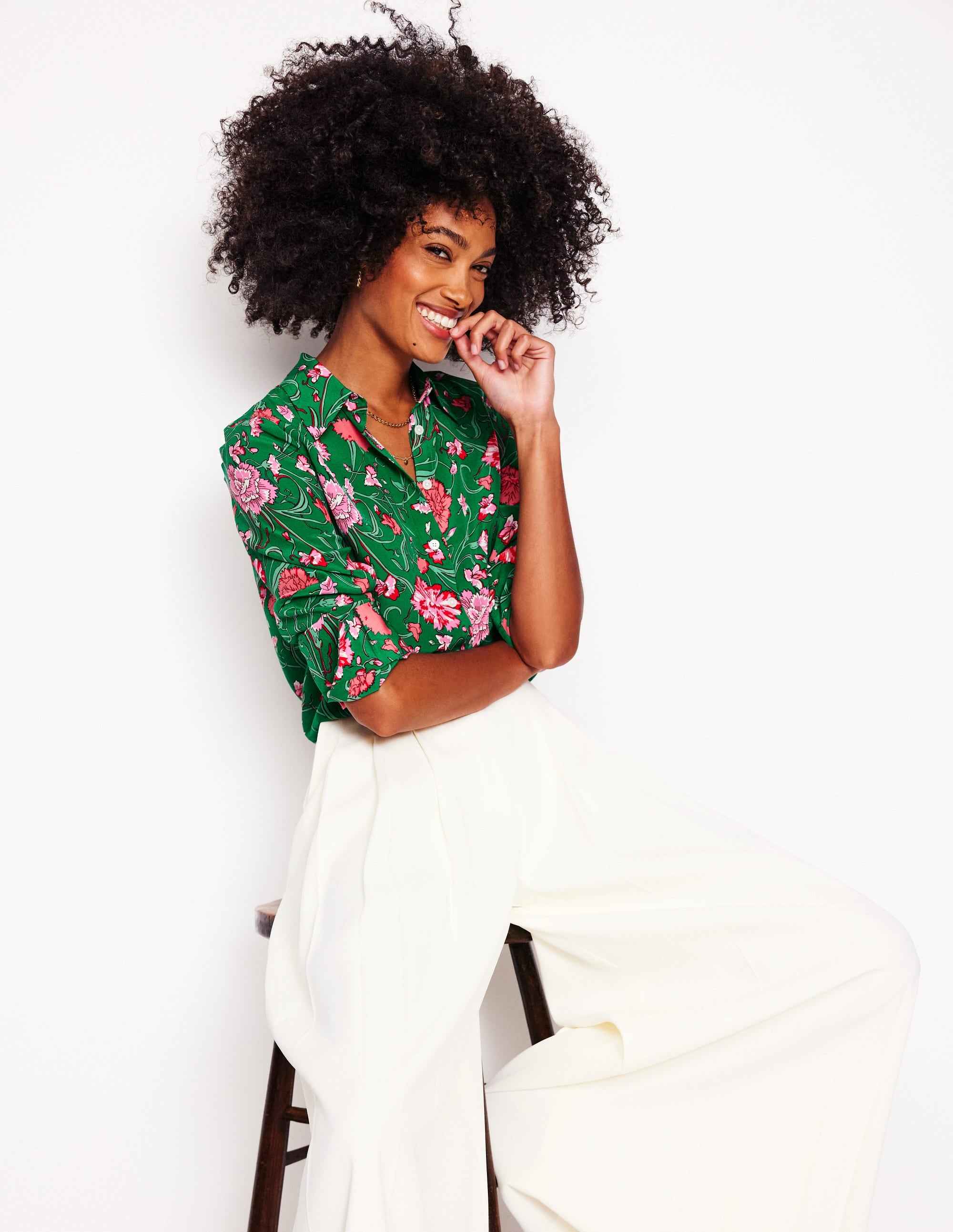 A woman wearing a green silk shirt with pink, red, and white floral prints, and white trousers, standing against a white background.