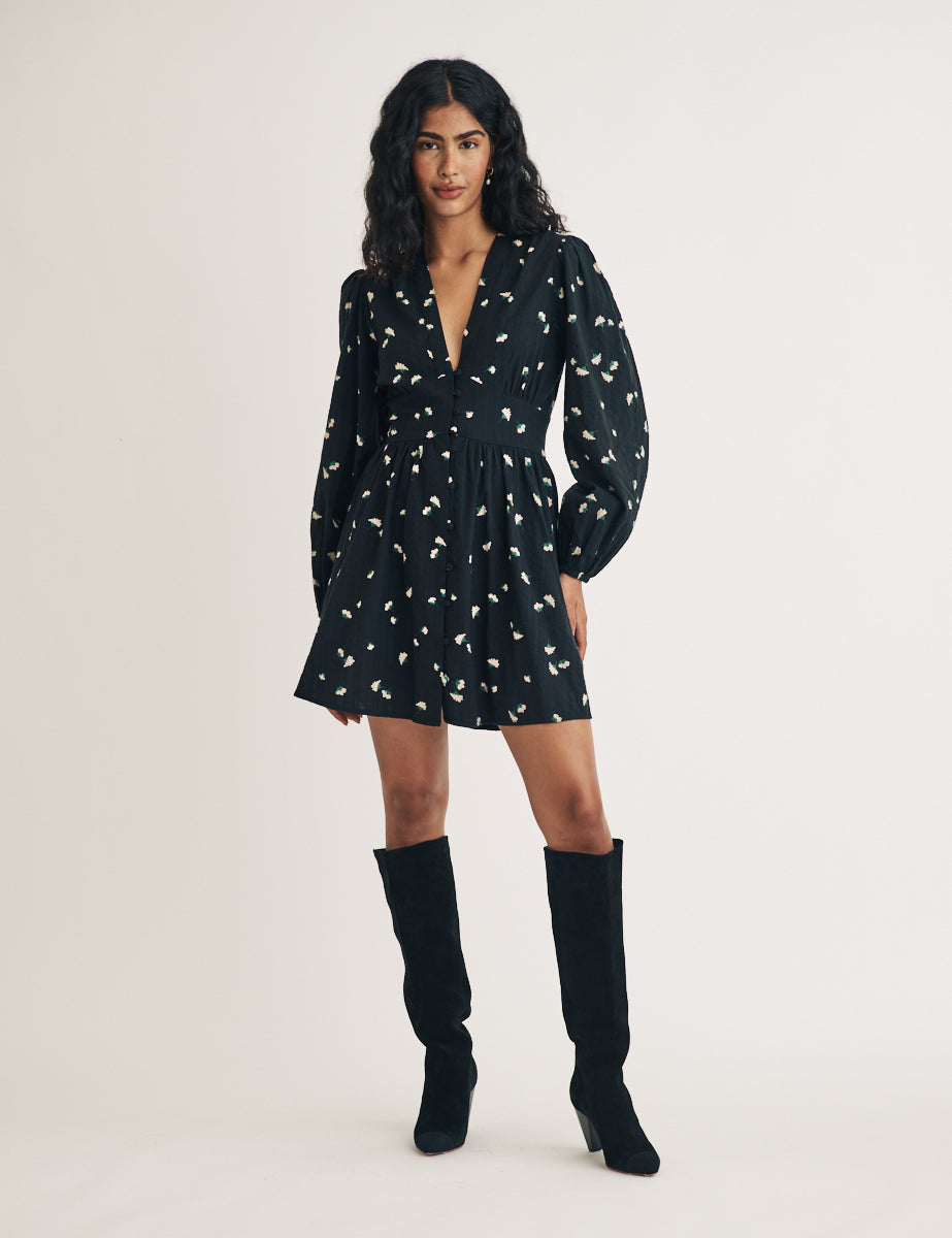 A woman wearing a black mini dress with a floral pattern, long balloon sleeves, and a V-neck, paired with black knee-high boots.