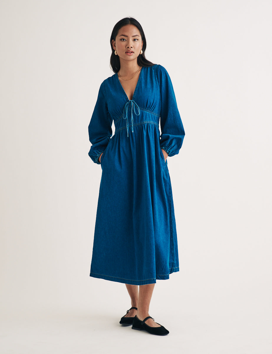 A woman wearing a blue denim midi dress with balloon sleeves, a V-neckline with tie detail, and an A-line skirt.