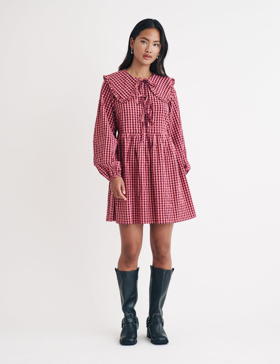 A woman wearing a pink and burgundy gingham mini dress with a Peter Pan collar, long puff sleeves, and a tie detail on the bodice.