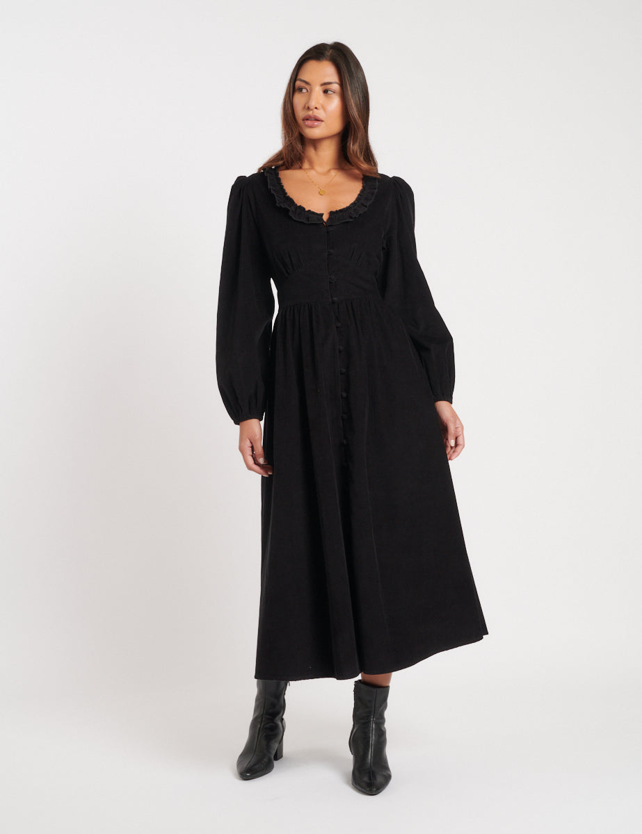 A woman wearing a black midi dress with long puff sleeves, a frilled collar, and a button-down front, paired with black ankle boots.