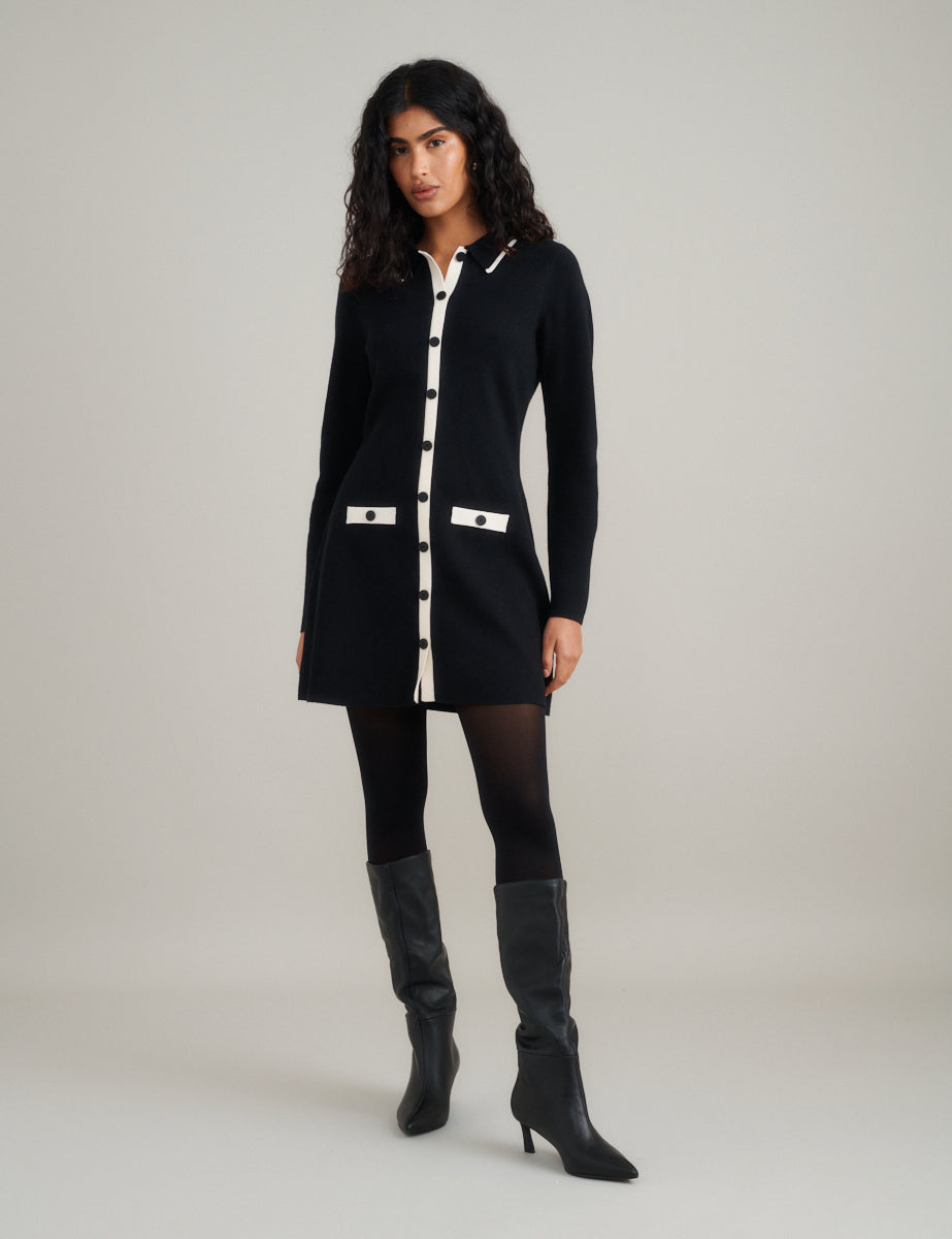A woman wearing a black knitted mini dress with white contrast trims, long sleeves, a preppy collar, and black buttons down the front, paired with black tights and knee-high black boots.