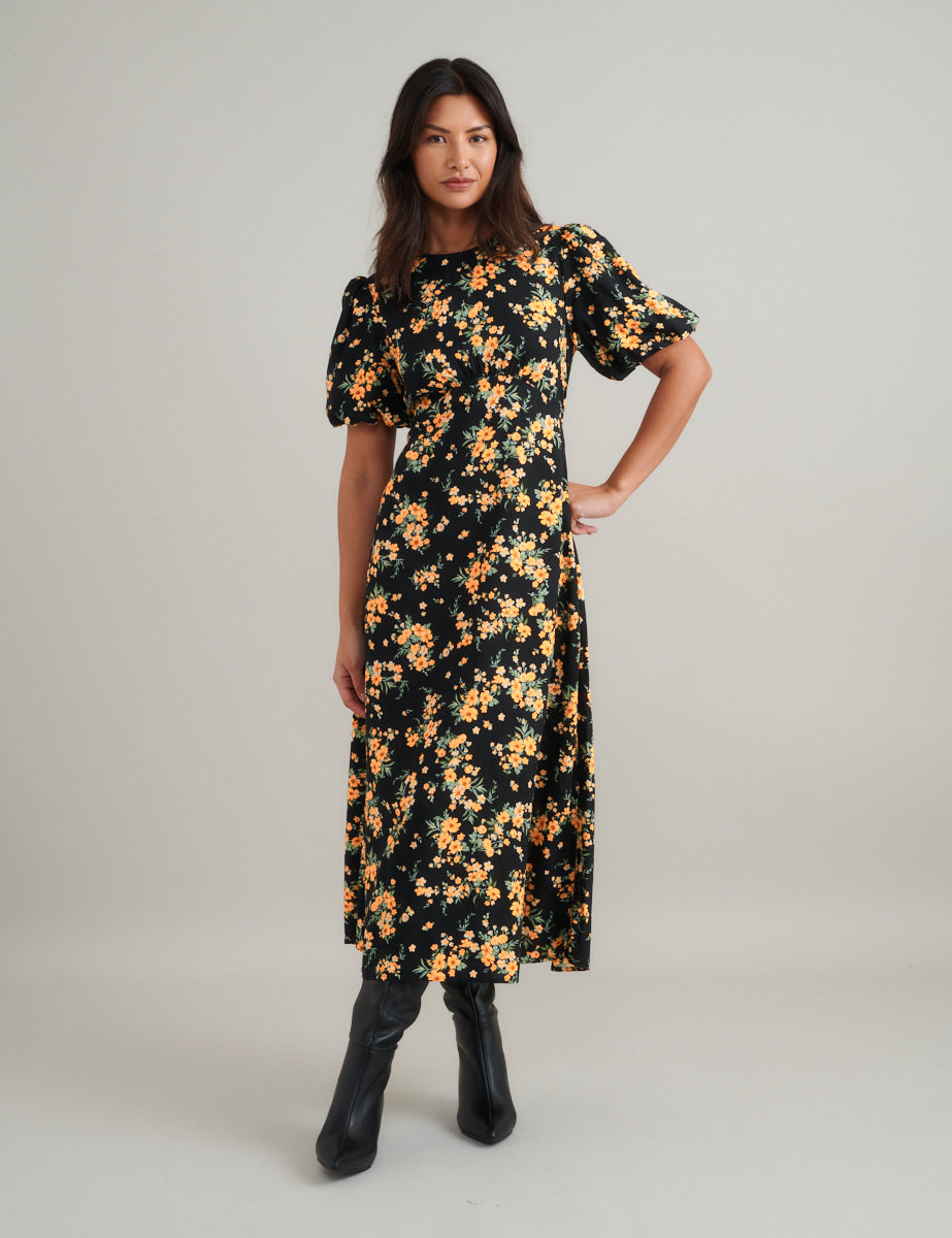 A woman wearing a black floral midi dress with puff sleeves and a curved waist seam, standing against a plain background.
