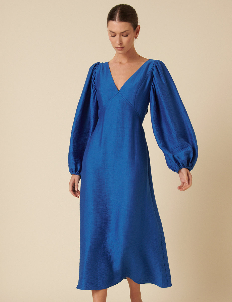 A woman wearing a vibrant blue midi dress with a square neckline, puffed balloon sleeves, and a backless design, standing against a plain background.