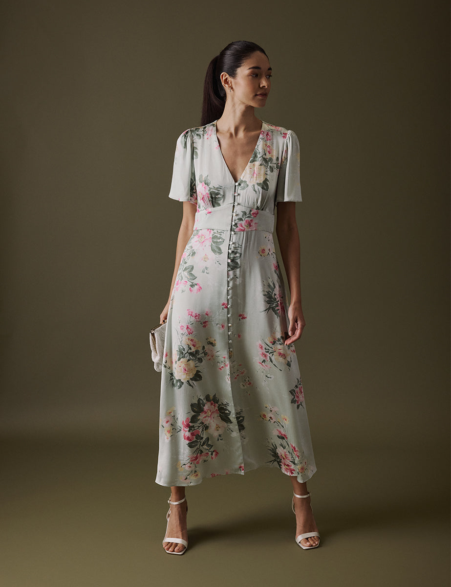 A woman wearing a floral green V-neck midi dress with fluted sleeves, a fitted waist, and a slit in the front, paired with white high-heeled sandals.