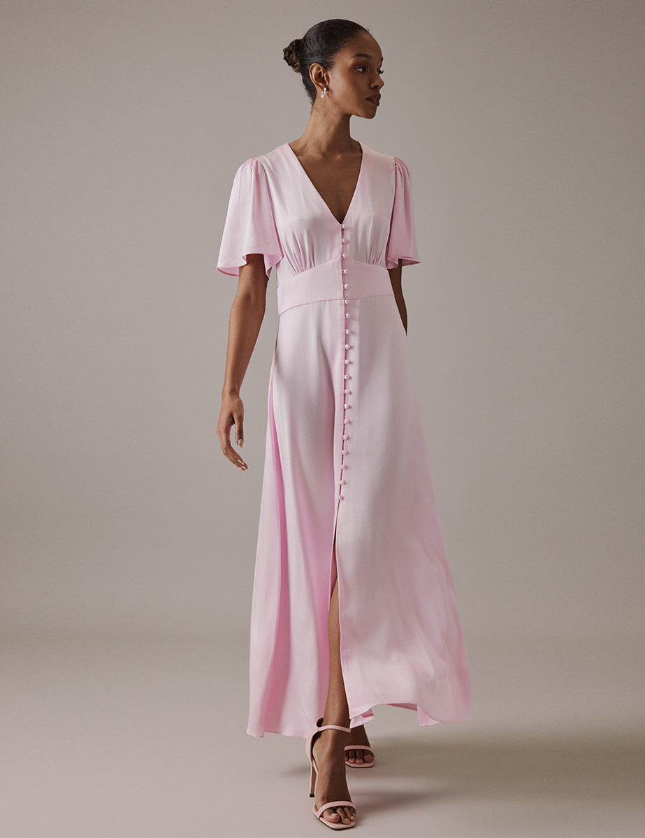 A woman wearing a soft pink satin midi dress with a V-neck, short fluted sleeves, and a slit in the front, styled with nude heels.