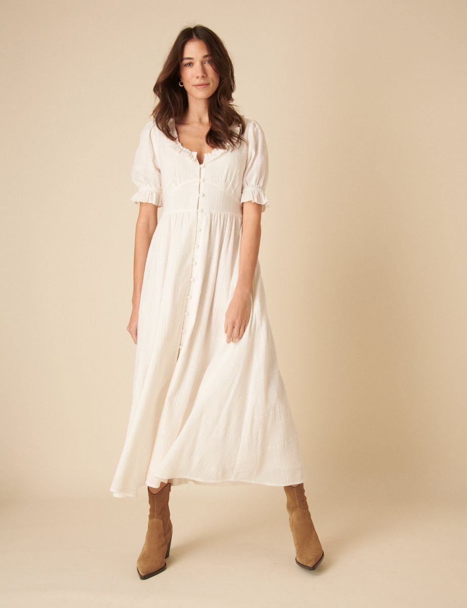 A woman wearing a white midi dress with a textured fabric, frilled collar, short puff sleeves, and a centre slit, standing against a plain background.