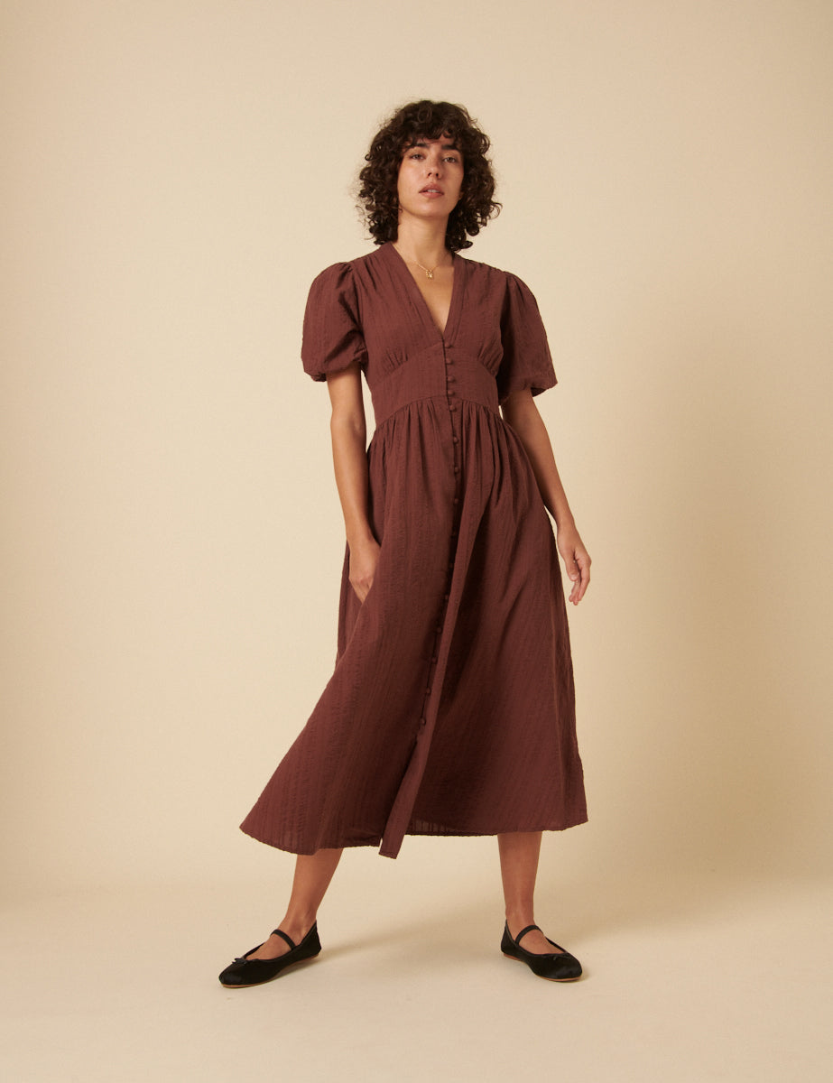 A woman wearing a brown midi dress with a V-neck, short puff sleeves, and an A-line skirt, standing against a plain background.