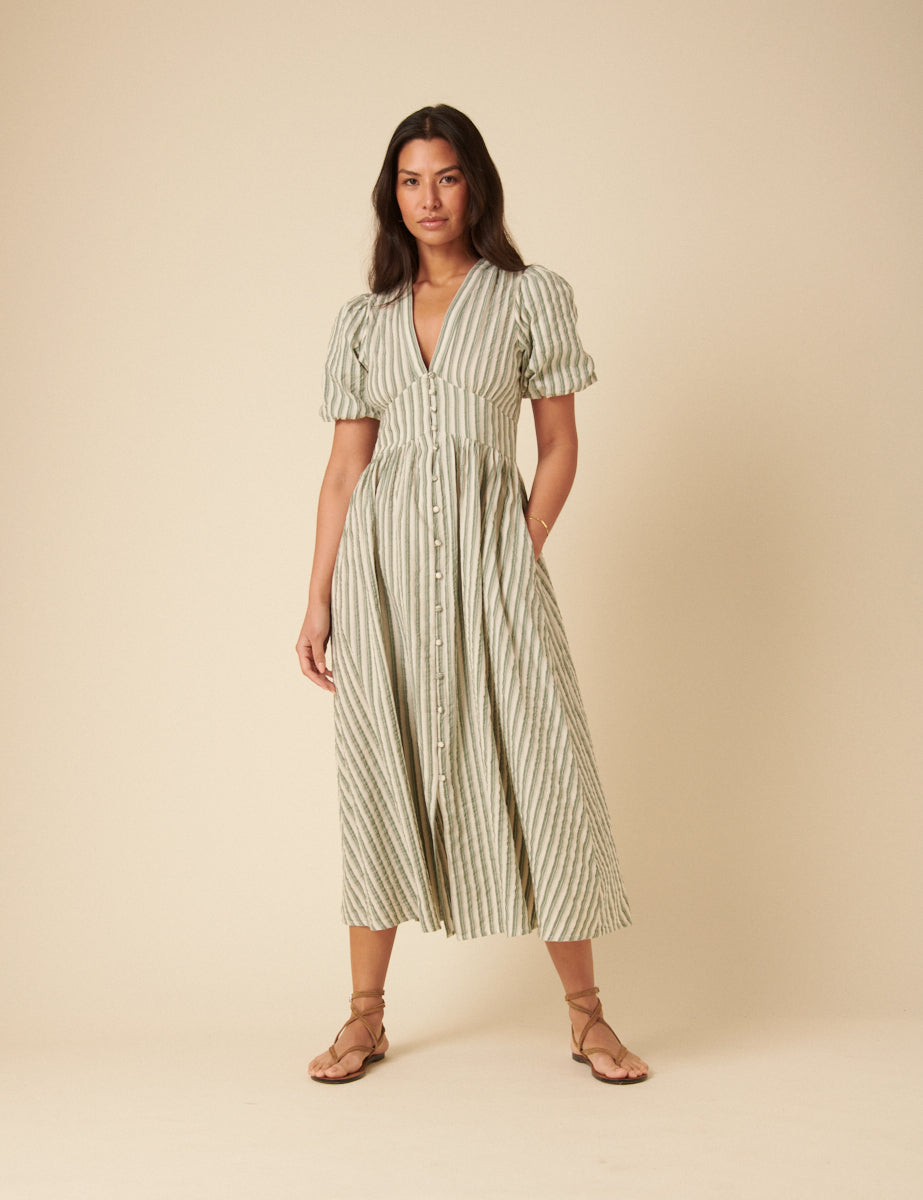 A woman wearing a green pinstriped midi dress with short puff sleeves, a V-neckline, covered buttons down the front, and a panelled waist, standing against a plain background.