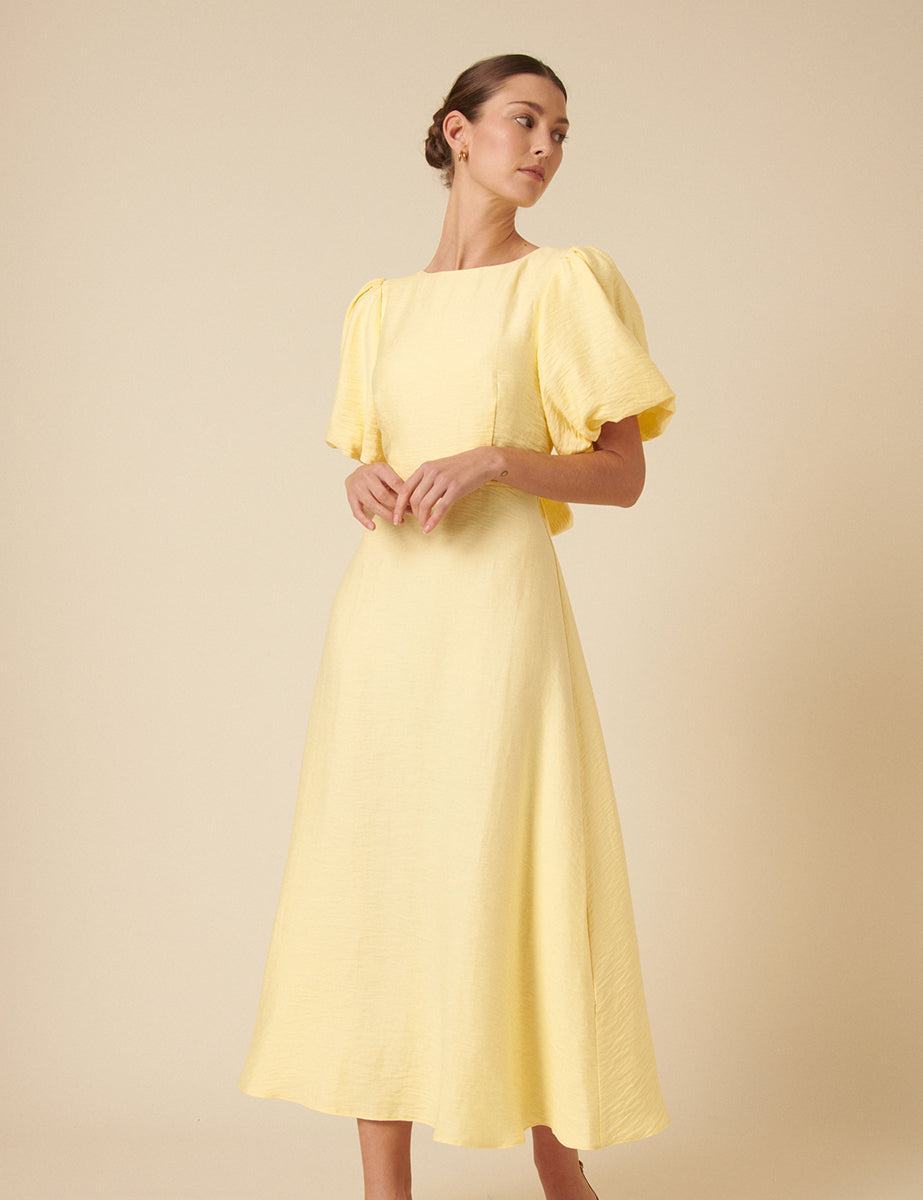 A woman wearing a yellow midi dress with puff sleeves, an open back with a bow, and a crew neckline.