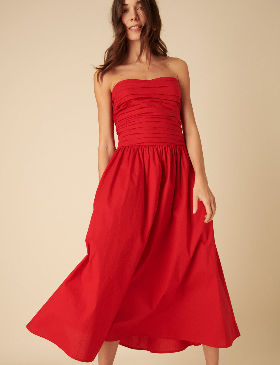 A woman wearing a bright red bandeau midi dress with a gathered bodice and a flowing skirt, standing against a plain background.