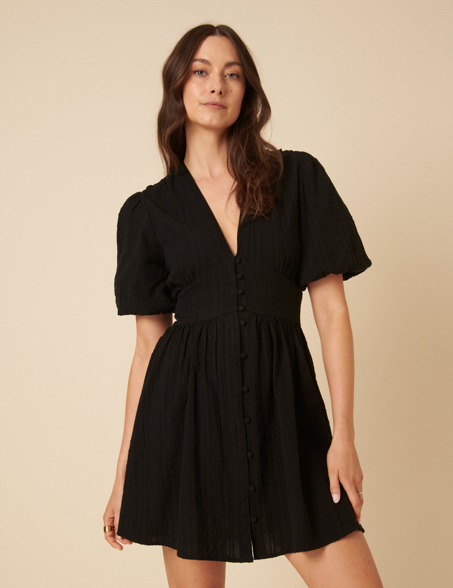 A woman wearing a black mini dress with a V-neck, puff sleeves, and buttons down the front, standing against a plain background.
