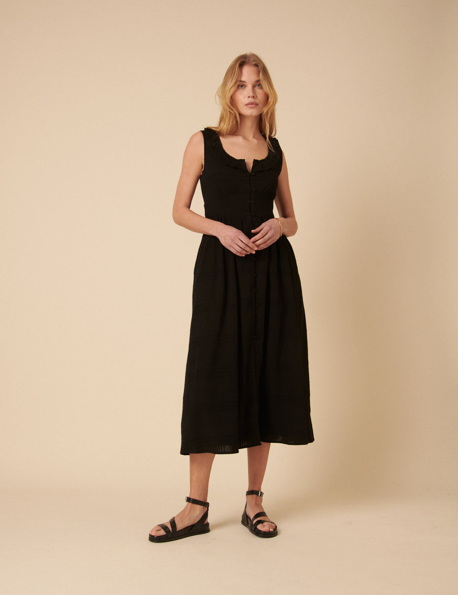 A woman wearing a black sleeveless midi dress with a round neckline, puff sleeves, and a panelled waist, paired with black sandals.