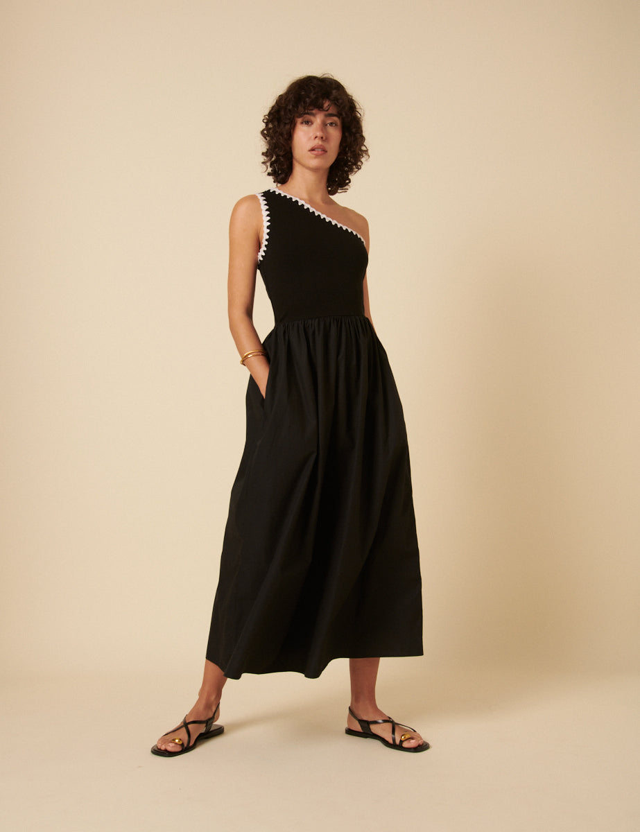 A woman wearing a black one-shoulder midi dress with crochet trim at the neckline, featuring an asymmetric design and flowing skirt.