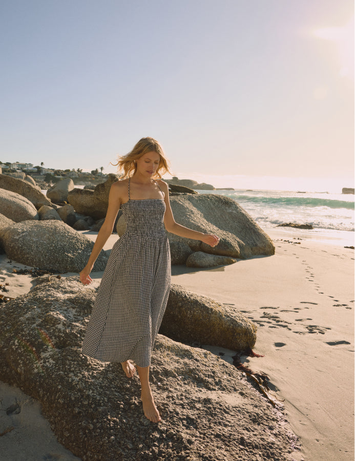 A woman wearing a black gingham midi dress with spaghetti straps and a square neckline, standing on rocks by the beach during sunset.