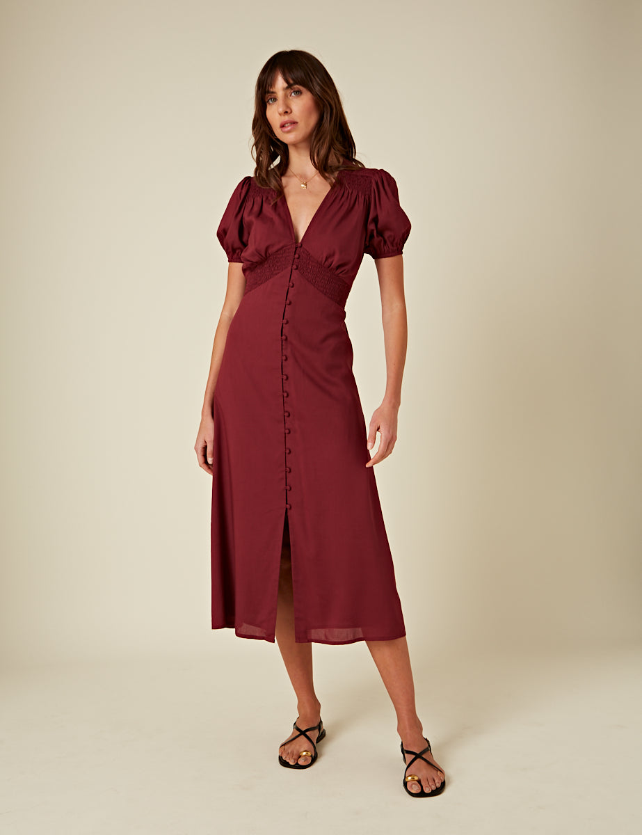 A woman wearing a burgundy satin midi dress with a V-neck, short puffed sleeves, and a button-down front, styled with black sandals.