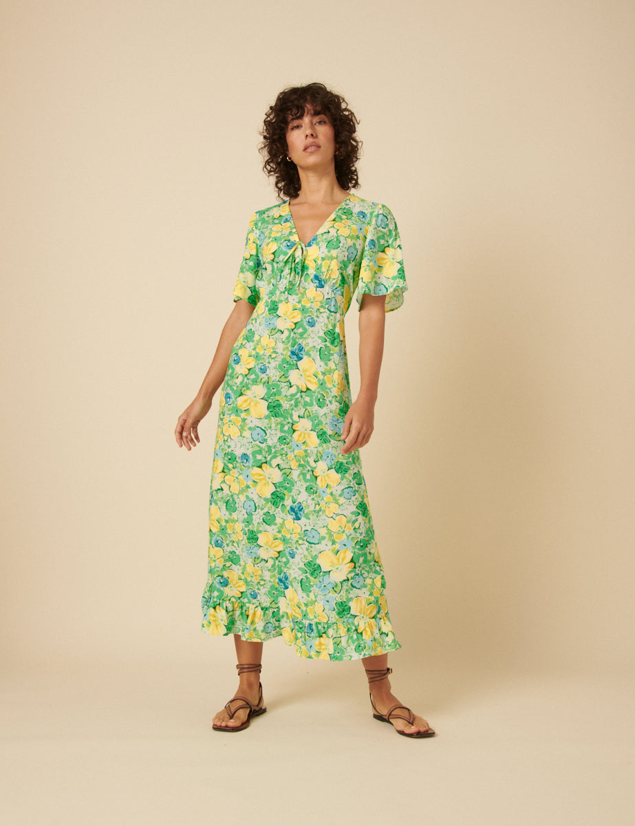 A woman wearing a green floral midi dress with yellow flowers, V-neckline with tie, short sleeves, and a relaxed skirt, standing against a plain background.