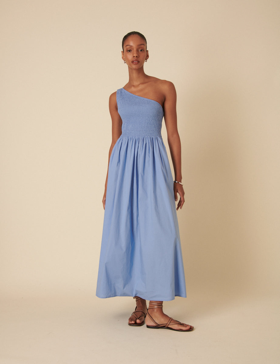 A woman wearing a pale blue, one-shoulder midi dress with an elasticated waist and an A-line skirt, standing against a plain background.