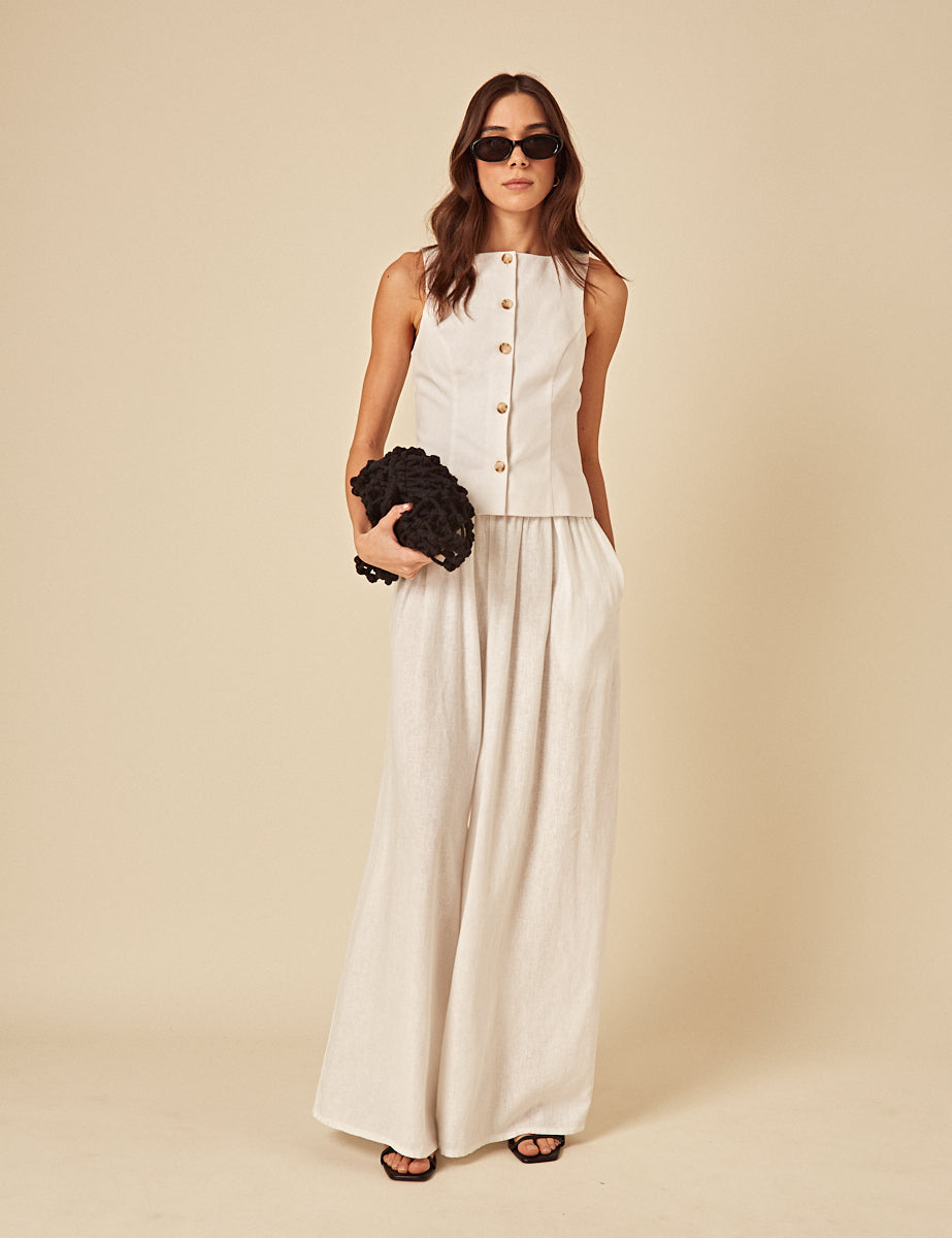 A woman wearing wide-leg, cream-colored linen-blend trousers with a pleated design, high waist, side pockets, and an elasticated back, paired with a sleeveless top and sunglasses.