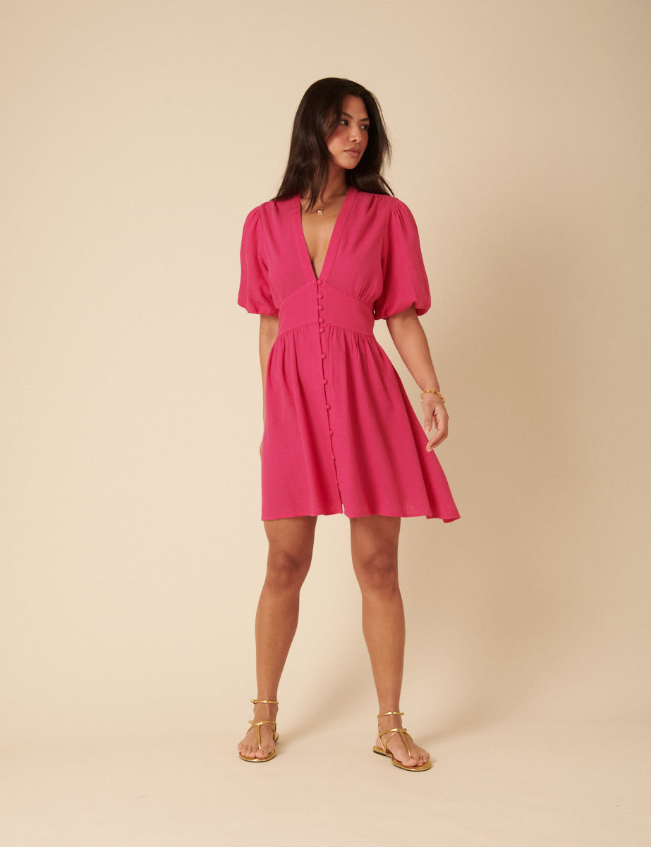 A woman wearing a vibrant pink mini dress with a V-neckline, short puff sleeves, and a button-down front, standing against a plain background.
