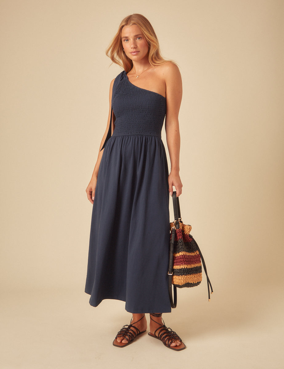 A woman wearing a navy one-shoulder midi dress with a shirred bodice, a bow at the shoulder, and a tiered A-line skirt, paired with black sandals and a multicolored woven bag.