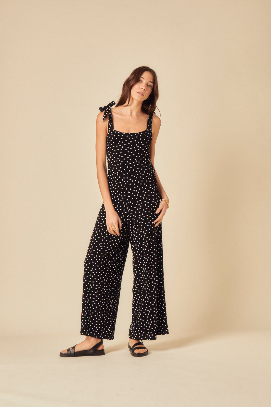 A woman wearing a black polka dot jumpsuit with a square neckline, frilly shoulder straps tied into bows, and wide-leg pants.