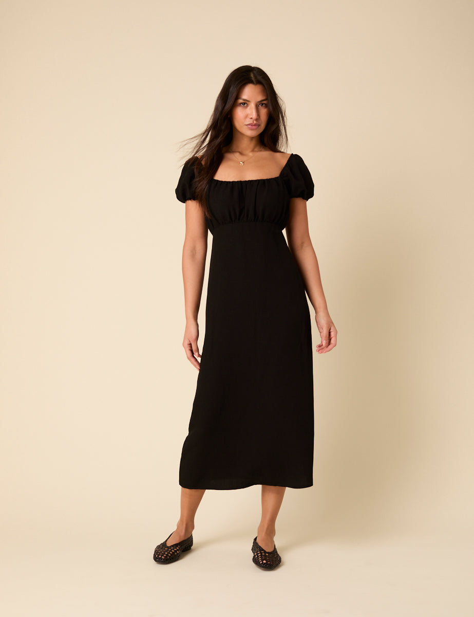 A woman wearing a black midi dress with puff sleeves and a ruched bodice, standing against a plain background.