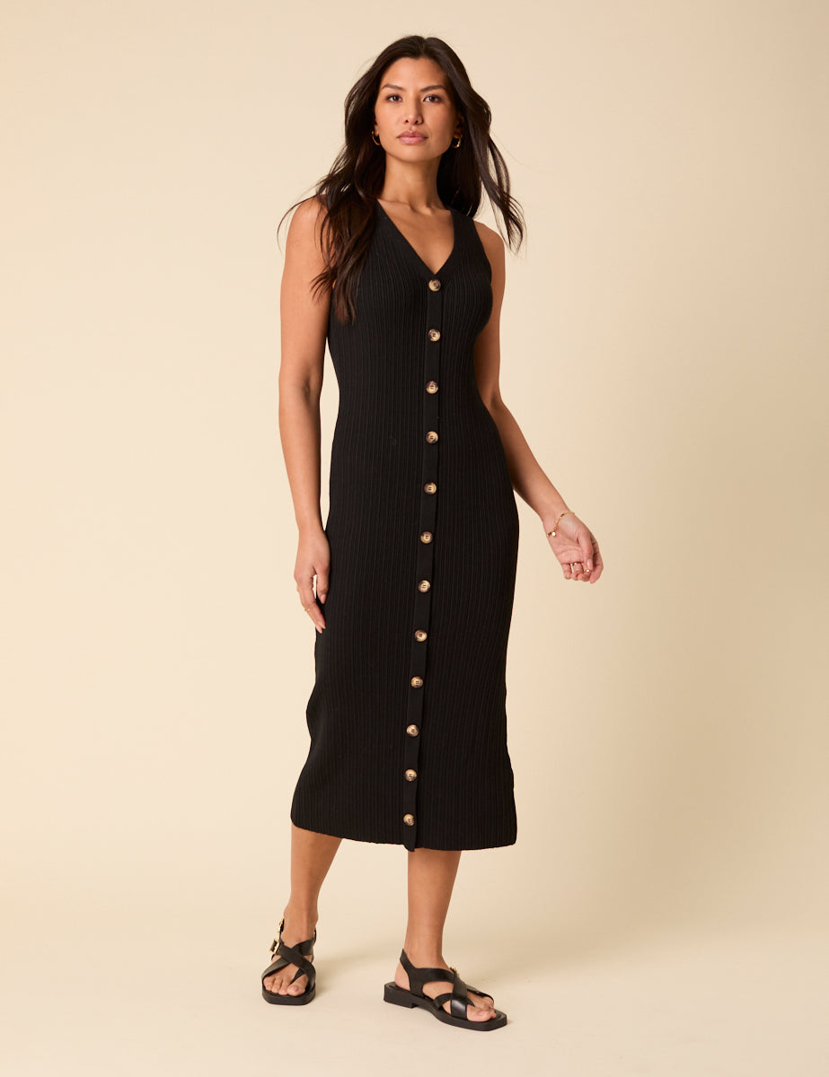 A woman wearing a black ribbed midi dress with a V-neckline and contrasting buttons down the front, paired with black sandals.
