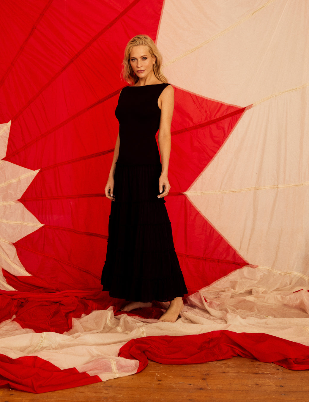 A woman wearing a black tiered midi dress with a fitted bodice, crew neckline, and thick straps, standing in front of a red and beige backdrop.