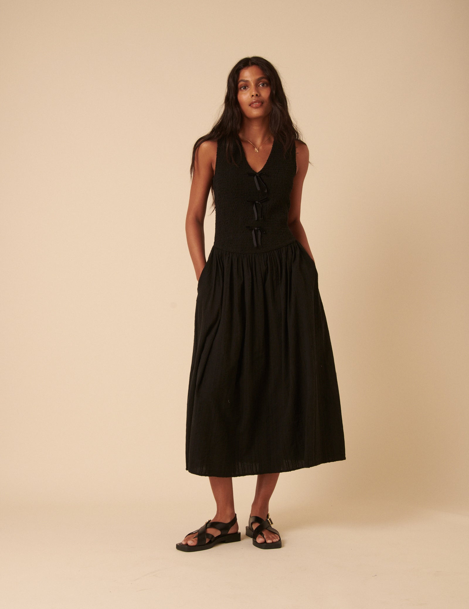 A woman wearing a black embroidered midi dress with a shirred bodice, bow ties, and a flowing skirt, paired with black sandals.
