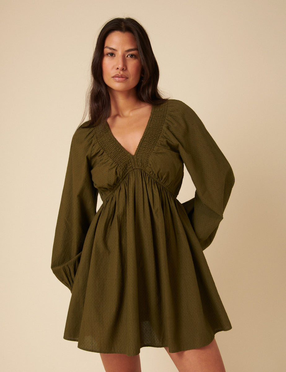 A woman wearing a khaki long sleeve mini dress with a V-neckline, puff sleeves, and a gathered waist.