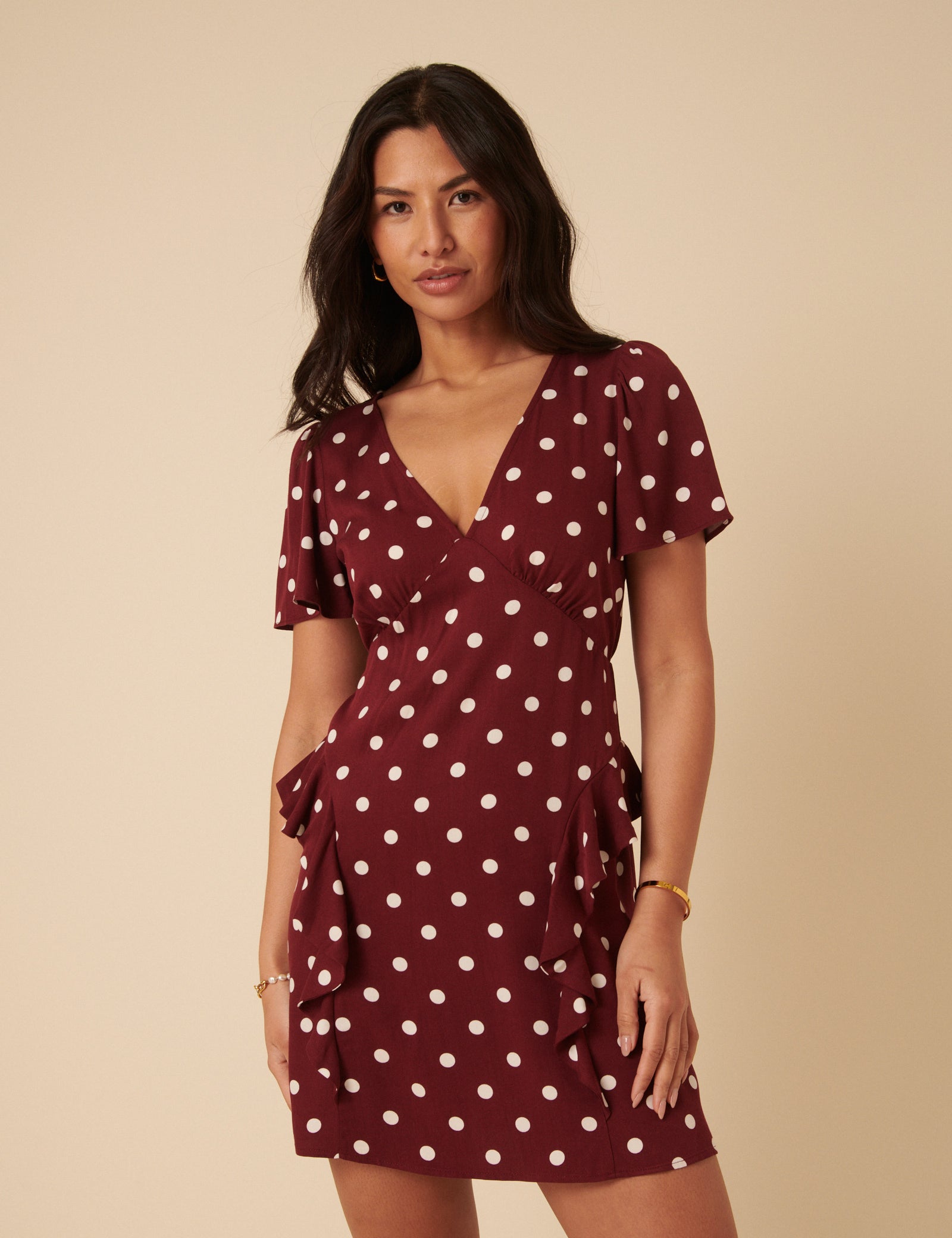A woman wearing a deep brown mini dress with white polka dots, V-neckline, flutter sleeves, ruffle trims, and a side slit.