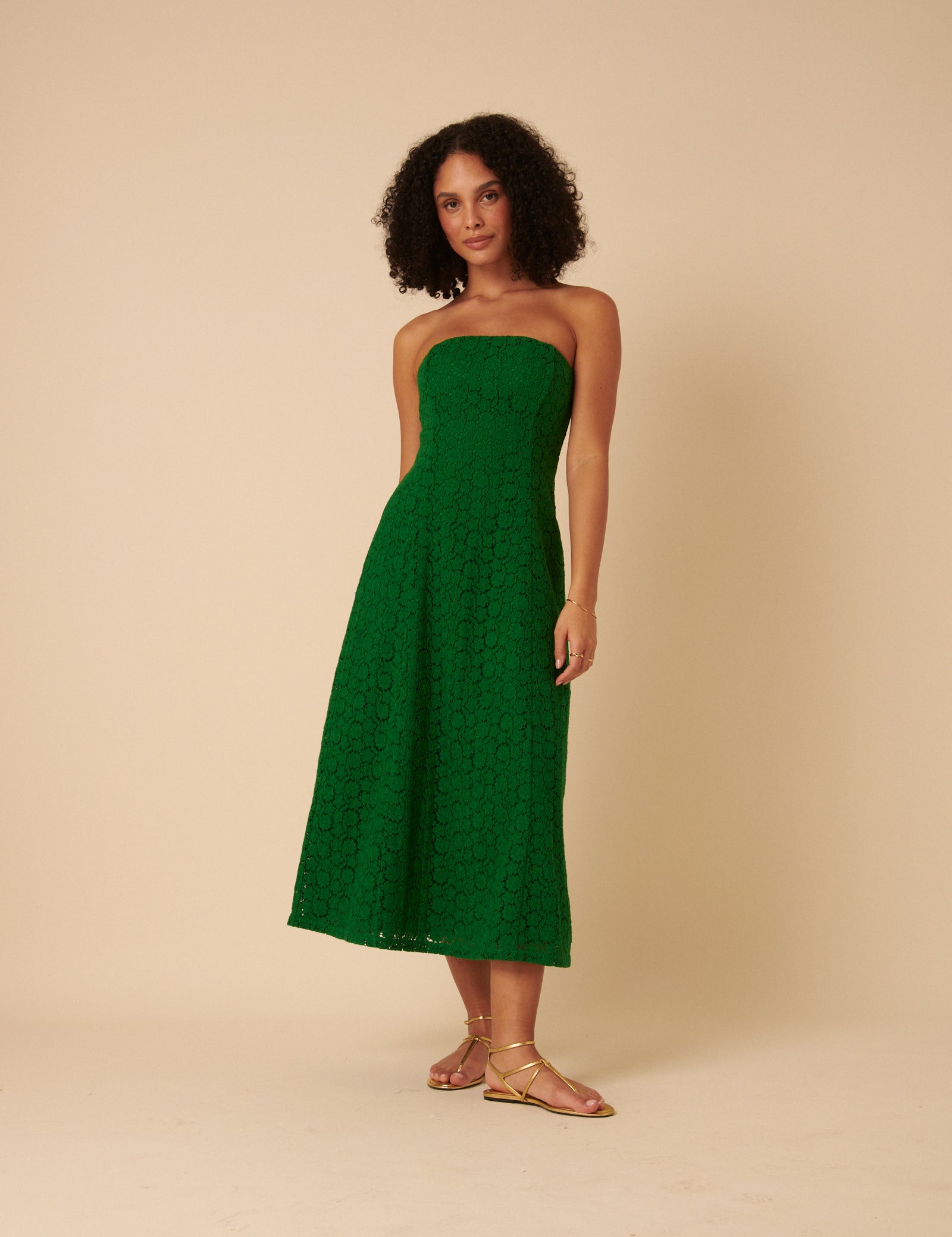 A woman wearing a green floral midi dress with a bandeau neckline and spaghetti straps, standing against a beige background.
