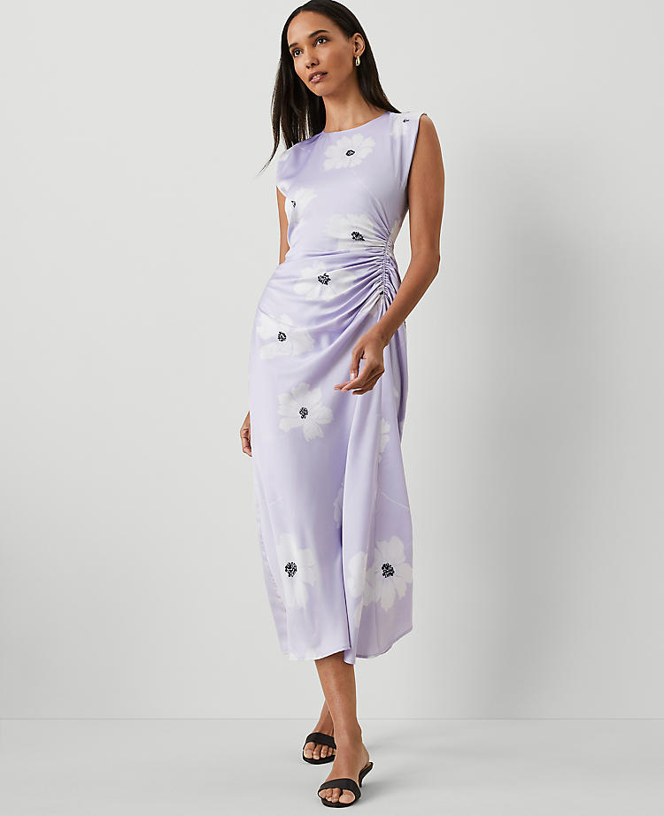 A woman wearing a light purple floral midi dress with gathered side drape, round neckline, cap flutter sleeves, and black open-toe heels.