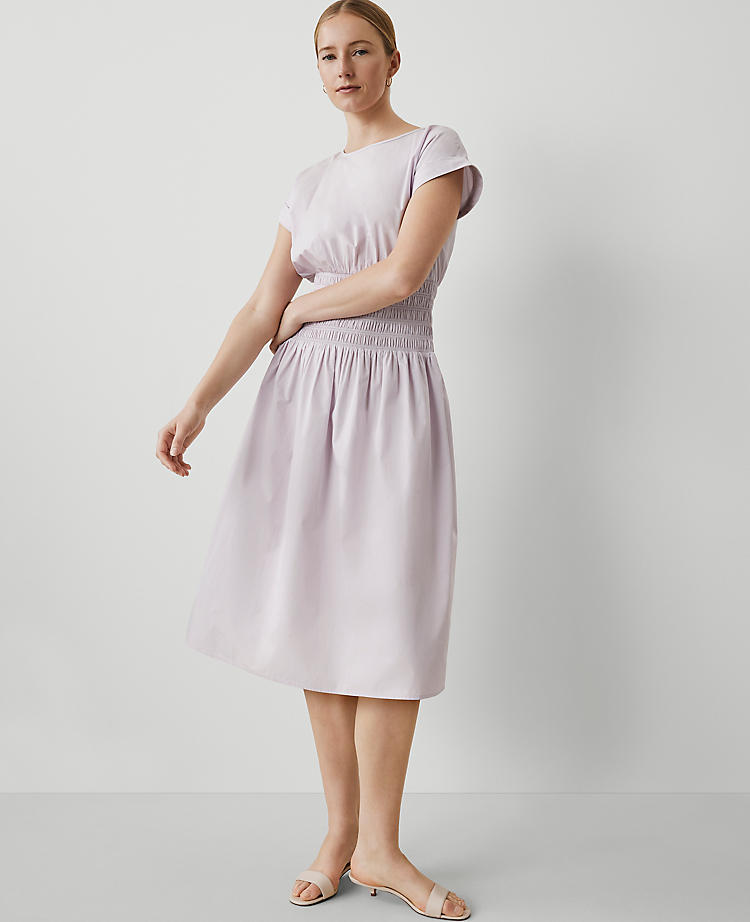 A woman wearing a light pink midi dress with a smocked waist, round neck, and cap sleeves, standing against a plain background.