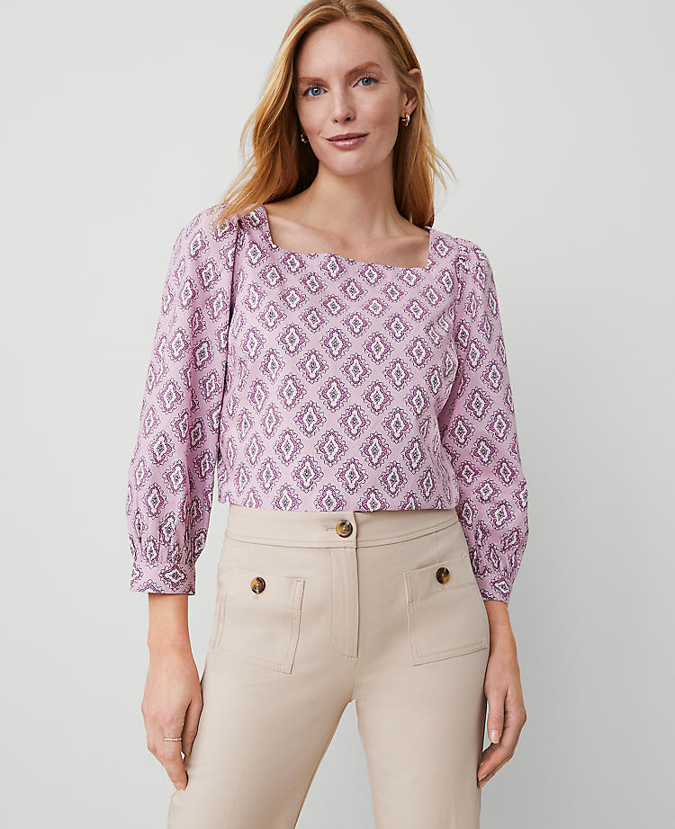 A woman wearing a pink and purple patterned blouse with a square neckline and puff sleeves, paired with beige pants.