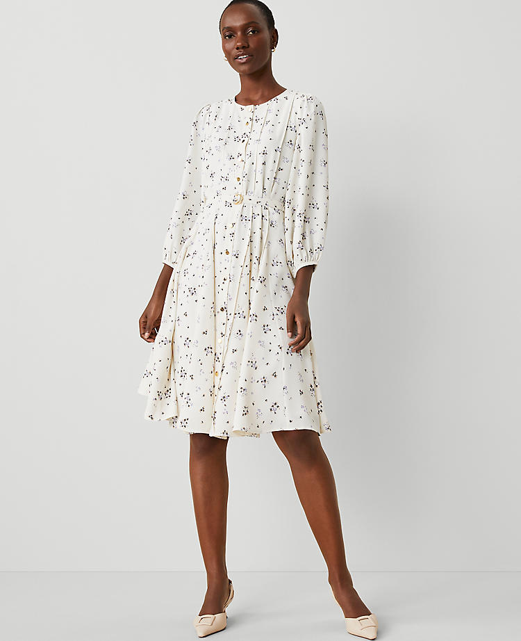 A woman wearing a cream-colored floral dress with seamed panels, puff shoulders, and a D-ring belt, standing against a plain background.